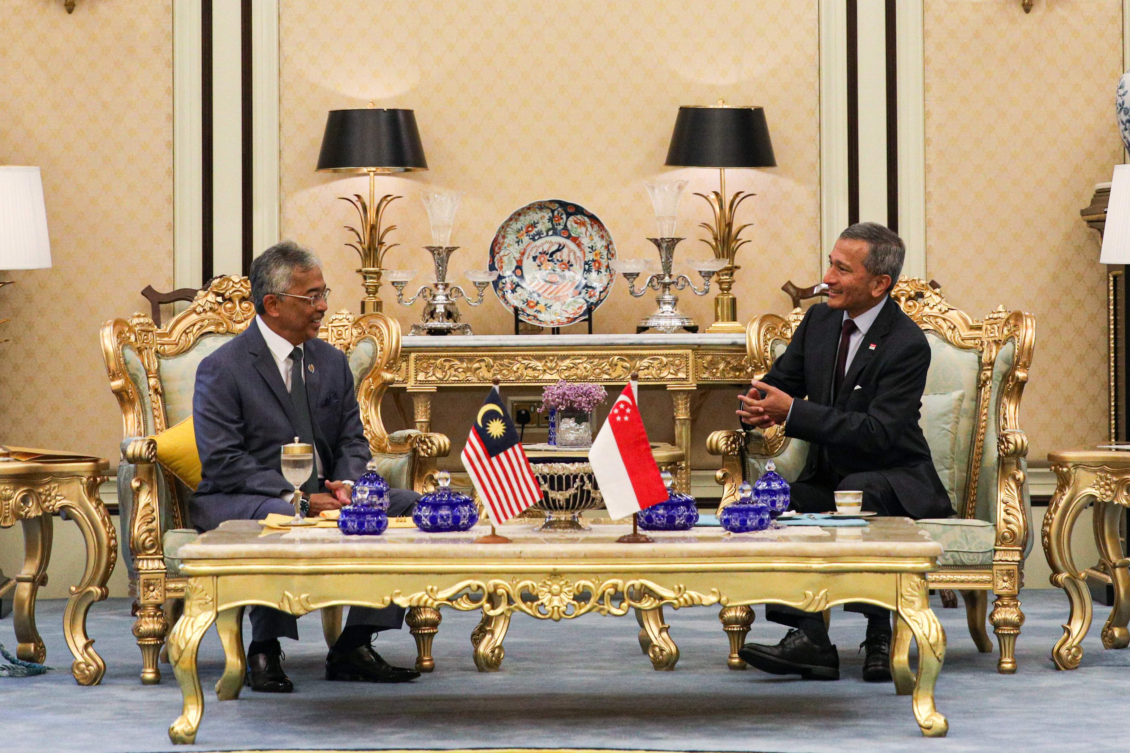 Two men in suits sit at a table with Malaysian and Singaporean flags.